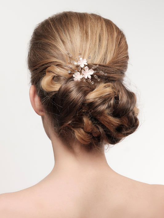 A bride's elegantly styled updo, featuring soft curls intricately woven together, adorned with delicate pink floral hairpins, providing a timeless and sophisticated look.