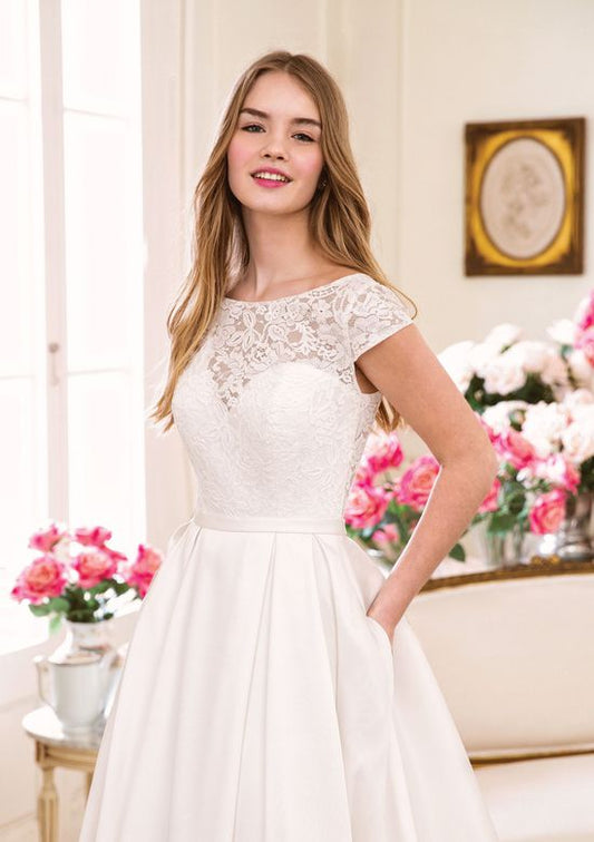 A close-up of a bride in a wedding gown with a lace bodice and cap sleeves, showcasing the delicate floral lacework and the elegant simplicity of the design. The soft pleats of the satin skirt and the bride's relaxed pose enhance the gown's timeless charm.