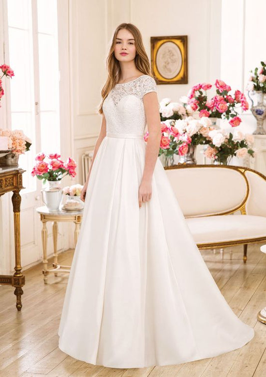 A bride wearing a classic A-line wedding gown with a lace bodice and cap sleeves. The gown features a high neckline and a full satin skirt that gracefully flows to the floor, creating an elegant and timeless look. The scene is set in a bright, floral-decorated room, enhancing the gown's romantic charm.